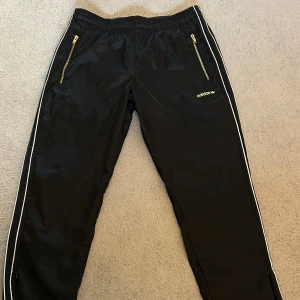 Adidas retro träningsbyxor - Svarta Adidas retro träningsbyxor med guldiga dragkedjor och logga. Byxorna har vita ränder längs sidorna och resår i midjan. Perfekta för sport eller avslappnad streetwear stil. Klassisk Adidas-design med raka ben och dragkedja vid benen. (De är använda endast ett handfull gånger)