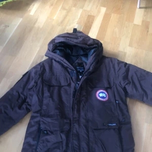 Brun dunjacka från Canada Goose - expedition parka - Säljer en brun dunjacka från Canada Goose med huva och klassisk logga på bröstet. Jackan har flera fickor framtill och dragkedja. Perfekt för kalla dagar när du vill hålla dig varm och samtidigt se snygg ut.