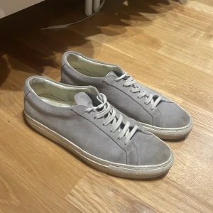 Common Projects - Säljer ett par stilrena grå sneakers från Common Projects i mjuk mocka. Skorna har vit platt sula, rund tå och klassisk snörning. Perfekta för dig som gillar minimalistisk design och hög kvalitet.
