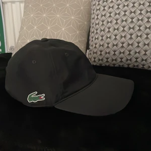 Svart keps från Lacoste Sport - Säljer en klassisk svart keps från Lacoste Sport med den ikoniska krokodilloggan broderad på sidan. Kepsen har böjd skärm och justerbar passform baktill. Perfekt accessoar för en sportig och stilren look. Keps o kofta