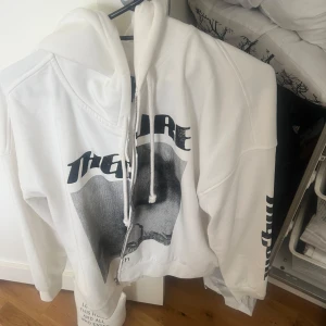 Vit hoodie med tryck - Säljer en vit hoodie med svart och grått tryck framtill och text på ena ärmen. Tröjan har huva med dragsko och en avslappnad passform. Perfekt för en chill och stilren look.