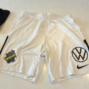 Vita AIK fotbollsshorts från Nike - Säljer ett par vita AIK fotbollsshorts från Nike med Dri-FIT-material. Shortsen har AIK:s klubbmärke på ena benet och Volkswagen-logga samt Nike Swoosh på det andra. Resår och snörning i midjan för skön passform.