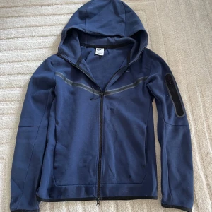 Marinblå Nike tech  - Snygg marinblå Nike tech från Nike med hel dragkedja och huva. Nike techen  har en diskret svart detalj på ärmen med dragkedja och en tunn reflexrand över bröstet. Perfekt för chill dagar ute eller när du vill ha en sportig look.