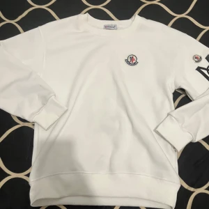 Moncler tröja - Säljer en stilren vit sweatshirt från Moncler med deras ikoniska logga på bröstet och ärmen. Tröjan har långa ärmar och ribbade muddar vid ärmslut och nederkant. Perfekt för en avslappnad och trendig look.