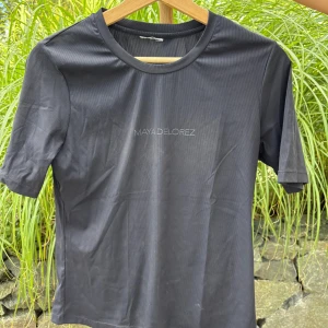 Svart t-shirt från Maya Delorez - Svart t-shirt från Maya Delorez med diskret logga på bröstet. Klassisk rund hals och korta ärmar. Materialet känns mjukt och har en lätt glans, perfekt för en clean och sportig look.