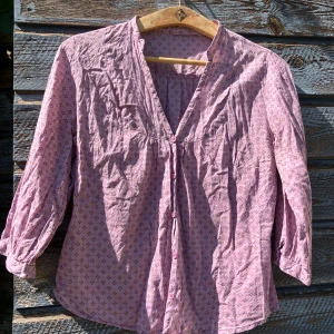 Ljuslila mönstrad skjorta med knappar - Söt ljuslila skjorta med små blommor och knappar framtill. Skjortan har en v-ringad hals och trekvartsärmar med knappslut. Materialet är mjukt och lätt, perfekt för vår och sommar. Passformen är normal och tyget har ett diskret mönster som ger en romantisk vibe.