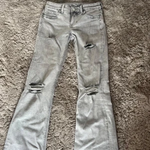 Grå bootcut jeans från H&M Divided - Snygga grå jeans med bootcut från H&M Divided, storlek 34. De har slitna detaljer med hål på knäna och låren för en cool vibe. Klassisk femficksmodell och knappgylf. Perfekta för dig som gillar streetstyle och vill sticka ut lite extra.