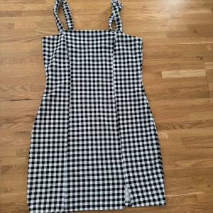 Svartvit rutig klänning H&M Divided - Supersnygg svartvit rutig klänning från H&M Divided i storlek L. Modellen är kort med axelband som knyts och har en liten slits framtill. Perfekt för dig som gillar en trendig och lekfull stil. Materialet är mjukt och känns bekvämt mot huden.