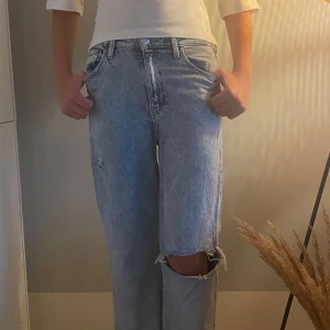 Ljusa wide jeans med hål på knät - Säljer ett par ljusblå jeans med breda ben och coolt hål på ena knät. Jeansen har klassisk femficksdesign, hög midja och raka bakfickor. Perfekt för dig som gillar avslappnad och trendig stil. Materialet är jeans och passformen är loose.