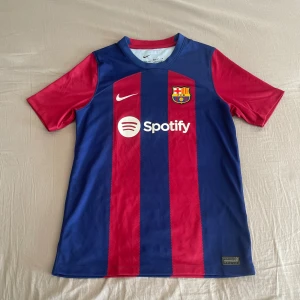 Barça matchtröja XL Nike Dri-Fit - Säljer en FC Barcelona matchtröja från Nike i storlek XL. Tröjan har klassiska breda blå och röda ränder, korta ärmar och rund hals. Materialet är lätt och andas, Dri-Fit polyester. Spotify-logga på bröstet och UNHCR-tryck på ryggen. Perfekt för dig som älskar Barça!