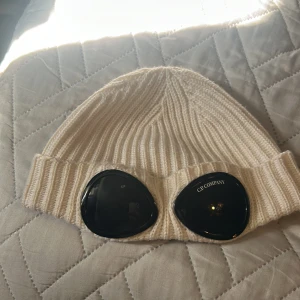 Vit ribbad mössa C.P. Company - Säljer en snygg vit ribbstickad mössa från C.P. Company med ikoniska svarta goggles framtill. Mössan har bred uppvikt kant och är tillverkad i mjukt stickat material som håller dig varm och ger en cool streetwear-look.