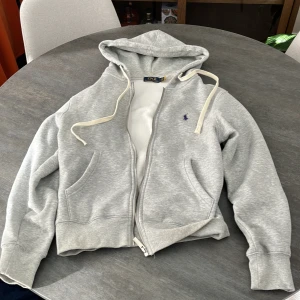 Grå hoodie från Polo Ralph Lauren - Säljer en grå hoodie från Polo Ralph Lauren med dragkedja och huva. Tröjan har vita snören, två fickor fram och ett litet blått Polo-logo broderat på bröstet. Mjuk insida och ribbade muddar vid ärmslut och nederkant. Perfekt för chill dagar. Pris kan diskuteras.