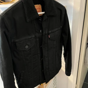 Svart fodrad jeansjacka  Levi's Sherpa - Säljer en svart jeansjacka från Levi's med mjuk fodrad Sherpa och krage. Jackan har klassiska bröstfickor med knappar, sidofickor och knappstängning framtill. Perfekt för dig som vill ha en cool och varm jacka med streetkänsla. Rek pris: 1499 kr