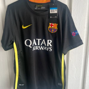 FC Barcelona Messi 10 svart matchtröja - Snygg FC Barcelona matchtröja från Nike med Messi 10-tryck på ryggen. Svart med gula detaljer och klubbmärke på bröstet, Champions League-patch på ärmen och Qatar Airways-logga framtill. Tillverkad i lätt Dri-FIT-material som andas. Perfekt för dig som älskar fotboll och Barca.