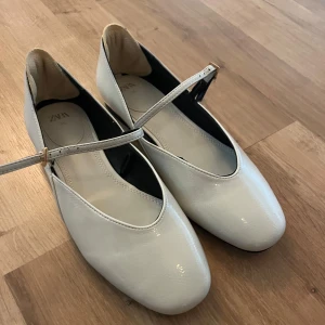 Vita ballerinaskor med rem från Zara - Säljer ett par stilrena vita ballerinaskor från Zara med rund tå och smal rem över foten som stängs med ett litet spänne i guld. Skorna har en platt sula och är gjorda i syntetmaterial med en lätt glansig finish. Perfekt för dig som gillar minimalistisk och chic stil.