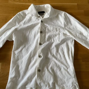 Vit manchesterskjorta - Snygg vit skjorta från Riley i mjuk manchester med tunna vertikala ränder. Klassisk krage, bröstficka och knappar framtill. Långärmad och perfekt för dig som gillar stilrena detaljer och skön passform. Skriv för frågor!