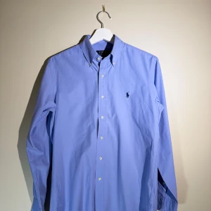 Blå skjorta från Polo Ralph Lauren - Snygg ljusblå skjorta från Polo Ralph Lauren med klassisk button-down krage och broderad logga på bröstet. Custom fit-modell med långa ärmar och vita knappar framtill. Perfekt för dig som gillar stilrena och tidlösa plagg.