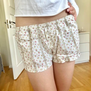 Blommiga shorts från Brandy Melville - Säljer ett par söta vita shorts från Brandy Melville med små rosa och gula blommor. Resten av shprtsen är krämvita. De har elastisk midja, knappar framtill och är gjorda i mjuk bomull. Passformen är mellan xs och s i passformen💛🩷