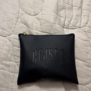 Svart kuvertväska från Kejsar - Snygg svart kuvertväska från Kejsar med präglad logga på framsidan. Väskan är tillverkad i skinnimitation och har en guldfärgad dragkedja upptill. Perfekt storlek för att bära med det viktigaste och passar till många olika stilar.