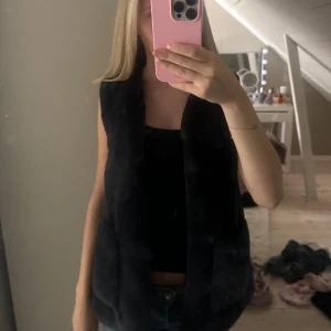 Marinblå pälsväst med bred krage - Mysig svart pälsväst med bred krage och öppen front. Västen är enkel och stilren, perfekt att slänga över en hoodie eller långärmad tröja. Den har en rak passform och känns riktigt fluffig och varm. Snygg att styla med jeans eller kjol för en trendig look.