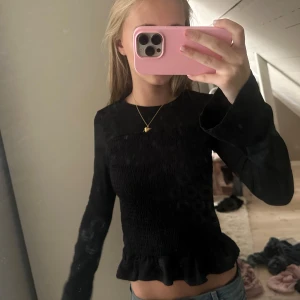 Svart långärmad topp med volang - Säljer en svart långärmad topp med smockad midja och volangkant nedtill. Modellen är croppad och har utsvängda ärmar som ger en snygg siluett. Perfekt att styla med jeans eller kjol för en trendig look.