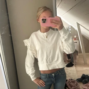 Vit croppad sweatshirt med volang - Säljer en vit croppad sweatshirt med volangdetalj på ena axeln. Tröjan har rund hals, långa ärmar och bred mudd nedtill. Perfekt för dig som gillar en clean look med lite extra edge. Materialet känns som mjuk bomull.