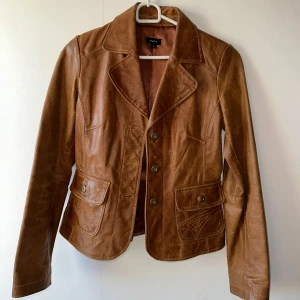 Brun skinnjacka från Lindex, strl 34 - Snygg brun skinnjacka från Lindex i klassisk kavajstil med krage och knappar framtill. Jackan har två fickor med lock och markerade sömmar. Perfekt för dig som gillar tidlös stil och vill ha något som sticker ut.
