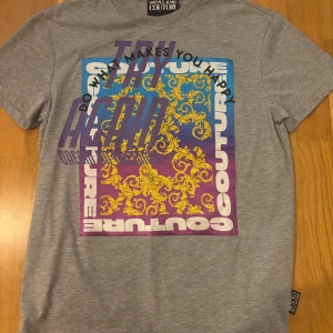 Grå Versace Jeans Couture t-shirt - Snygg grå t-shirt från Versace Jeans Couture i storlek M med korta ärmar. Framsidan har ett stort tryck med färgglada barockdetaljer i gult, blått och lila samt texten 'TRY AGAIN'. Baksidan har coolt rött och svart tryck med ord som 'EPIC' och 'STRONG'. Tillverkad i mjuk bomull.