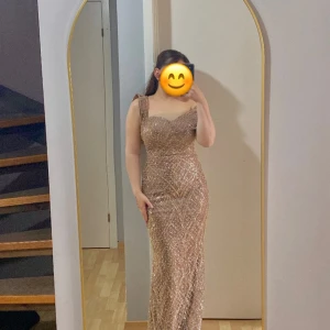 Glittrig champagnefärgad långklänning - Säljer en elegant långklänning i champagnefärg med glittrigt mönster och pärldetaljer. Klänningen har breda axelband, figurnära passform och ett snyggt sweetheart-urrringning. Perfekt för bal eller fest! Materialet känns lyxigt och skimrande.