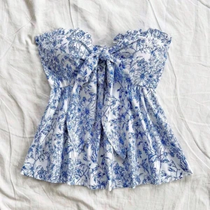 Blommig vit och blå bandeau topp - Supersöt bandeau topp med blått blommigt mönster på vit botten. Toppen har volangkant upptill, knytning framtill och smockad rygg för extra komfort. Luftig och flowy passform, perfekt för varma dagar. Materialet känns mjukt och lätt mot huden, endast testad💞