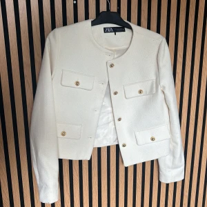 Vit kavajjacka från Zara - Snygg vit kavajjacka från Zara med rund hals och guldfärgade knappar. Jackan har fyra fickor framtill och är croppad i modellen. Perfekt för dig som gillar stilrena och klassiska plagg med en modern twist.
