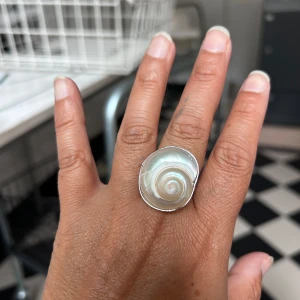 snäckring i silverfärg - Unik ring med en spiralformad snäcka i vit och pärlemor, infattad i silverfärgad metall. Ringen har en cool bohemisk vibe och sticker verkligen ut med sitt naturinspirerade snäckskal som centerpiece. Perfekt för dig som gillar statement-smycken och strandkänsla.