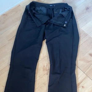 Svarta kostymbyxor från Bik Bok - Svarta kostymbyxor från Bik Bok i klassisk rak modell med midwaist. Byxorna har bälteshällor, diskreta bakfickor och stängs med dragkedja och knapp framtill. Materialet är mjukt och har en lätt glansig finish som ger en snygg look.