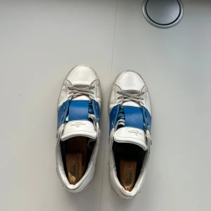 Valentino Open sneakers vit/blå - Säljer ett par Valentino Open sneakers i vitt skinn med bred blå panel över mitten. Skorna har rund tå, snörning och platt sula med mönstrad undersida. Snygga nitar på sidan av sulan ger extra edge. Perfekta för dig som gillar lyxiga street vibes. Nypris 7200 mitt pris 2900. Skick 6,5/10. Obs skorna tillverkas inte längre