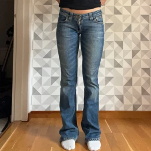 Blå bootcut jeans från Nudie Jeans - Säljer ett par snygga blå bootcut jeans från Nudie Jeans i modellen Bootcut Eddie. Jeansen har klassisk femficksdesign, låg midja och coola slitningar framtill och baktill. Materialet är mjukt jeans med orangea sömmar och detaljer på bakfickorna. Storlek 27/34. 
