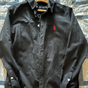Svart skjorta från Ralph Lauren M  - Svart skjorta från Ralph Lauren i klassisk passform med vit knappstängning och rött broderat logotyp på bröstet. Skjortan har lång ärm och är tillverkad i mjuk bomull. Perfekt för dig som gillar stilrena och tidlösa plagg. Det fattas två knappar som man kan se på bild 3 mellan de vita knapparna
