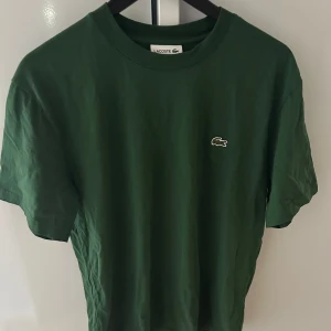 Grön t-shirt från Lacoste - Snygg grön t-shirt från Lacoste med klassisk krokodillogga broderad på bröstet. Modellen har rund hals och korta ärmar, perfekt för en chill och stilren look. Tillverkad i mjuk bomull som känns skön mot huden.