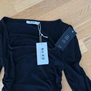 Svart långärmad klänning från NA-KD - Snygg svart långärmad klänning från NA-KD med rynkade detaljer längs sidorna som ger en cool och figurnära siluett. Klänningen har en rundad halsringning och är gjord i mjukt, stretchigt material som sitter skönt mot kroppen. Perfekt för dig som gillar stilrena och trendiga plagg.