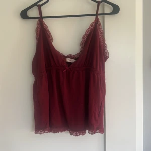 Vinröd linnetopp med spets från Nelly - Supersöt vinröd linnetopp från Nelly med tunna axelband och v-ringad hals. Spetsdetaljer längs kanterna och en liten rosett framtill ger en romantisk vibe. Mjukt material som känns skönt mot huden, perfekt för att lyfta din outfit.