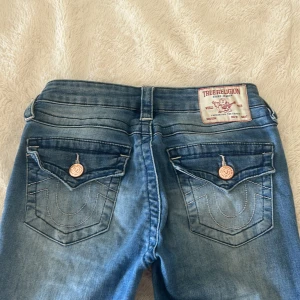 True Religion blå bootcut jeans  - Säljer ett par snygga blå jeans från True Religion med bootcut passform som ej går att köpa längre. De har coola detaljer som kopparfärgade knappar och fickor med broderade sömmar bak. Jeansen har låg midja och klassisk femficksdesign. Perfekta för dig som gillar en avslappnad men trendig stil. Dom är som nya! Inte använda mycket! Dom