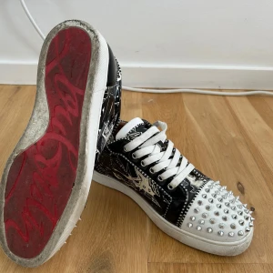 Christian Louboutin sneakers med nitar - Säljer ett par svarta och vita Christian Louboutin sneakers med coolt grafiskt mönster och silverfärgade nitar på tån. Skorna har klassisk röd sula, vita snören och detaljer på sidorna. Skitbra skick men har använts en del, medföljer enbart dustbag. Köpta här så oklart med äkthet därav pris