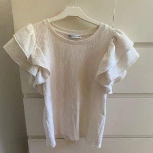Ribbad vit blus med volangärm från Zara - Supersöt ribbad blus från Zara i ljusvit ton med breda volangärmar som ger en extra touch. Rund halsringning och loose passform, perfekt för dig som gillar stilrena detaljer. Materialet är mjuk bomull och toppen har en lätt struktur som gör den unik. Storleken är lite liten i sig men tröjan är väldigt stretchig så den töjer ut sig efter en användning till din passform! 