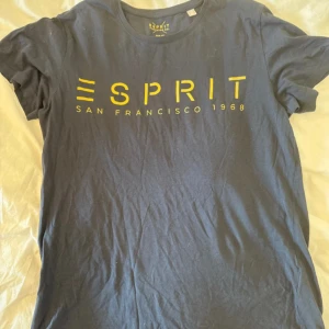 Blå t-shirt från Esprit med tryck - Snygg marinblå t-shirt från Esprit med stort gult tryck 'ESPRIT SAN FRANCISCO 1968' på bröstet. Klassisk rund hals och korta ärmar. Perfekt för dig som gillar enkel stil med coola detaljer. Gjord i mjuk bomull för skön känsla hela dagen.