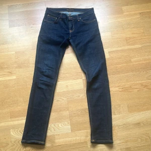 Nudie Jeans Long John mörkblå 29/32 - Snygga mörkblå jeans från Nudie Jeans, modell Long John. Slimmad passform med klassisk femficksdesign och orangea sömmar. Tillverkade i 98% ekologisk bomull och 2% elastan för extra stretch. Perfekt för dig som gillar en smal siluett och stilren look.