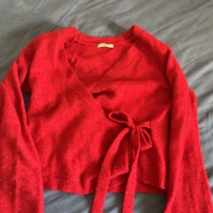 Röd omlottkofta från Gina Tricot - Säljer en snygg röd omlottkofta från Gina Tricot med långa ärmar och knytband i midjan. Koftan är stickad och har en v-ringad design som ger en chill och trendig vibe. Perfekt att styla med jeans eller kjol för en cool look.