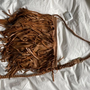 Fransväska i fejkläder - Säljer en brun axelväska i fejkskinn med långa fransar och flätat axelband. Älskar insidans fina tyg! Får plats med både dator och böcker💕💕 sliten i kanten därav priset