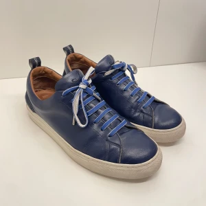  Finskor från Jacob Cohen - Marinblå sneakers från Jacob Cohen med vit och blå snörning, rund tå och vit platt sula. Skorna är tillverkade i mjukt skinn och i Italien. Vänster sko är lite sliten bak men inget man tänker på.