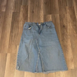 Ljusblå jeanskjol från Zara - Säljer en ljusblå jeanskjol från Zara med klassisk femficksdesign, elastisk midja och rå kant nedtill. Kjolen har en snygg slits framtill och bak, vilket ger en cool och avslappnad vibe. Perfekt för dig som gillar denim och vill ha en enkel men trendig look.