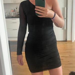 Svart oneshoulder kortklänning - Säljer en svart kortklänning med oneshoulder-design och långärmad meshärm. Klänningen är tight och har snygga rynkade detaljer längs sidorna som ger en cool siluett. Perfekt för dig som gillar en stilren look med lite edge.