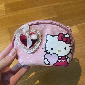 Hello Kitty rosa necessär med rosett - Söt rosa necessär med Hello Kitty-tryck, hjärtdetalj och rosett framtill. Perfekt för smink eller småprylar. Designen har vita prickar, hjärtan och en glansig kant. Hjärtdetaljen är dekorerad med spets och satinrosett för extra gullig look.